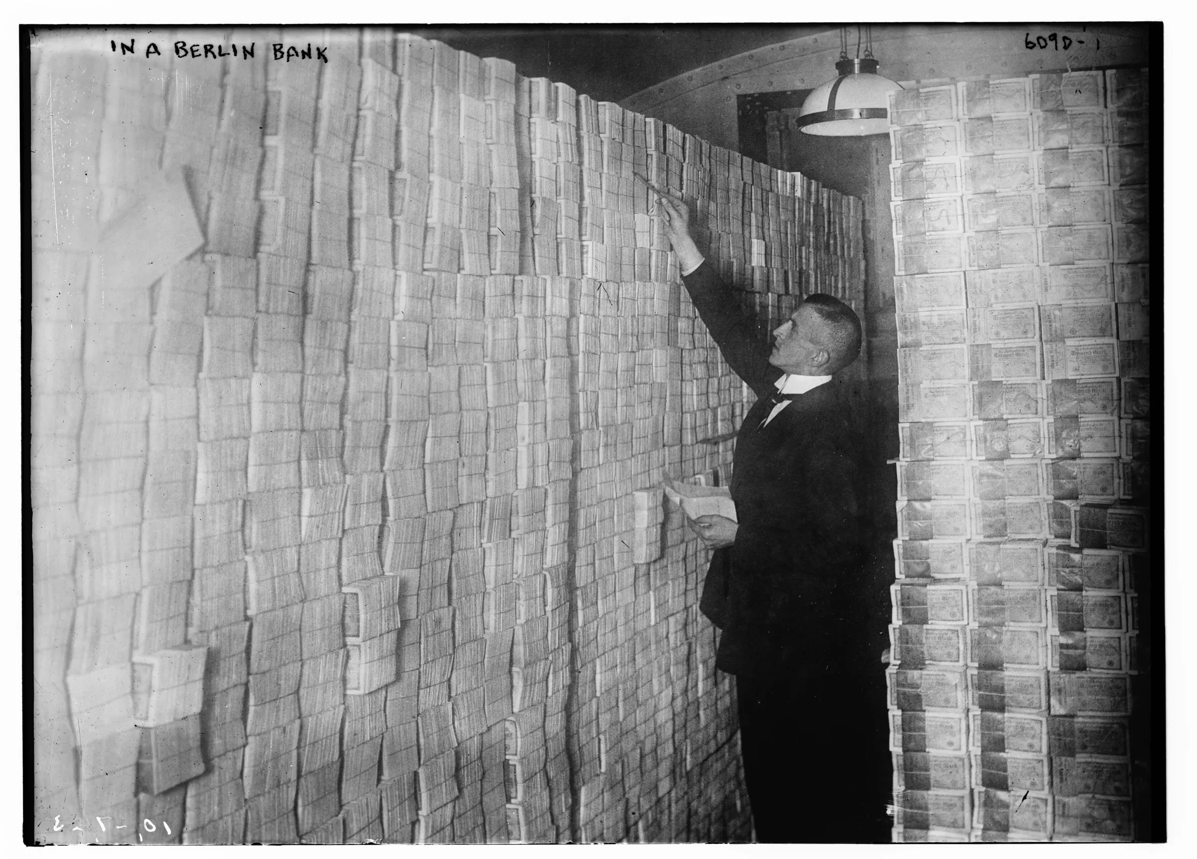 A man in a suit reaches up to a floor-to-ceiling wall of stacked banknote bundles in a bank vault lit by a single hanging lamp
