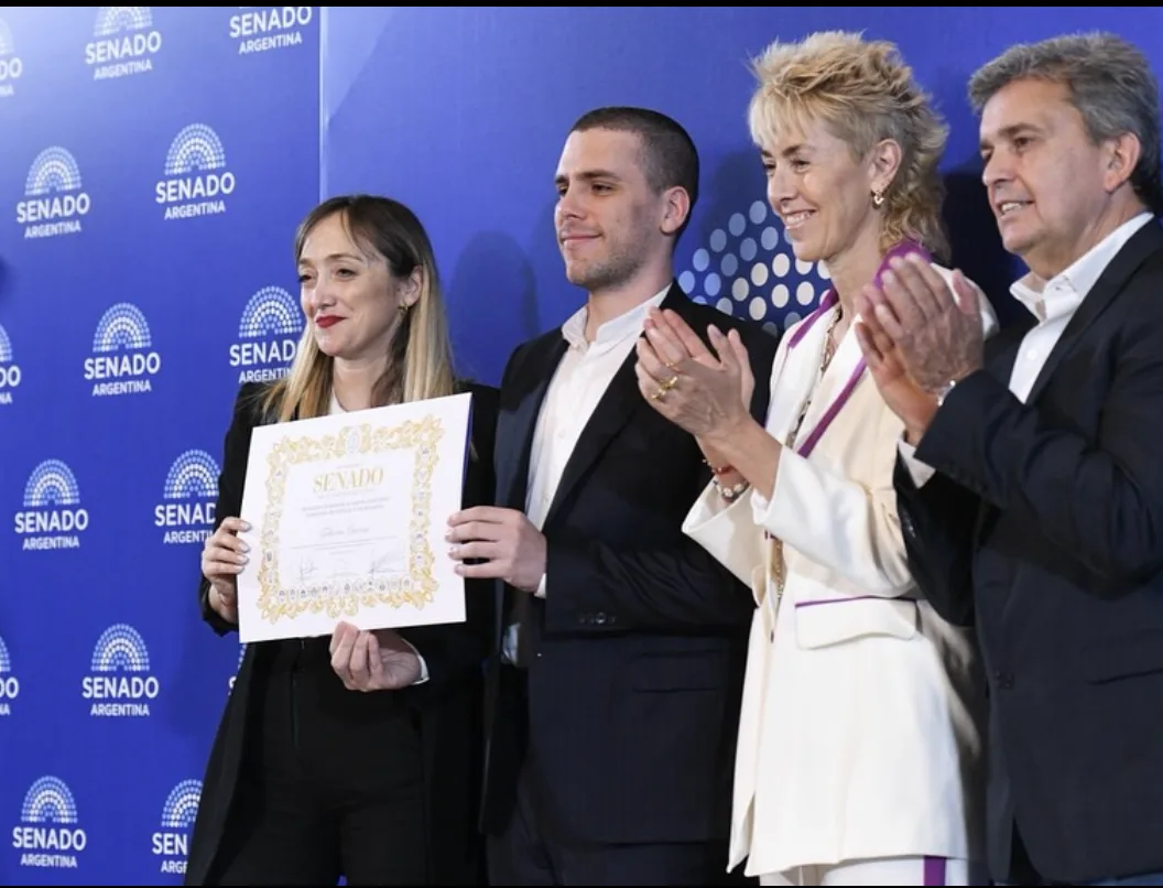 Four people standing in front of a blue SENADO Argentina backdrop, one holding up the Senate diploma while the others applaud
