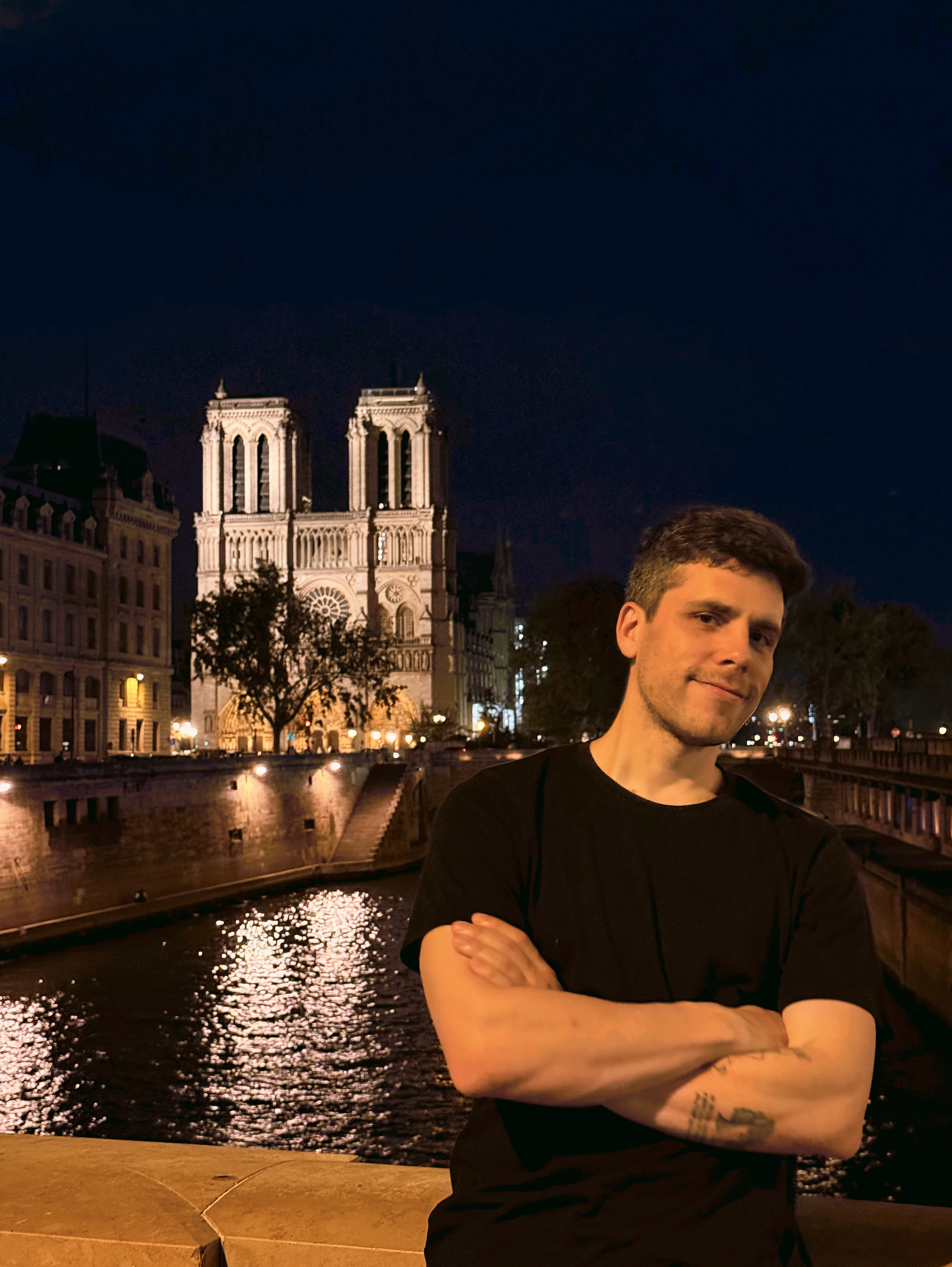Federico Carrone de noche en un puente sobre el Sena, de brazos cruzados, con Notre-Dame iluminada detrás