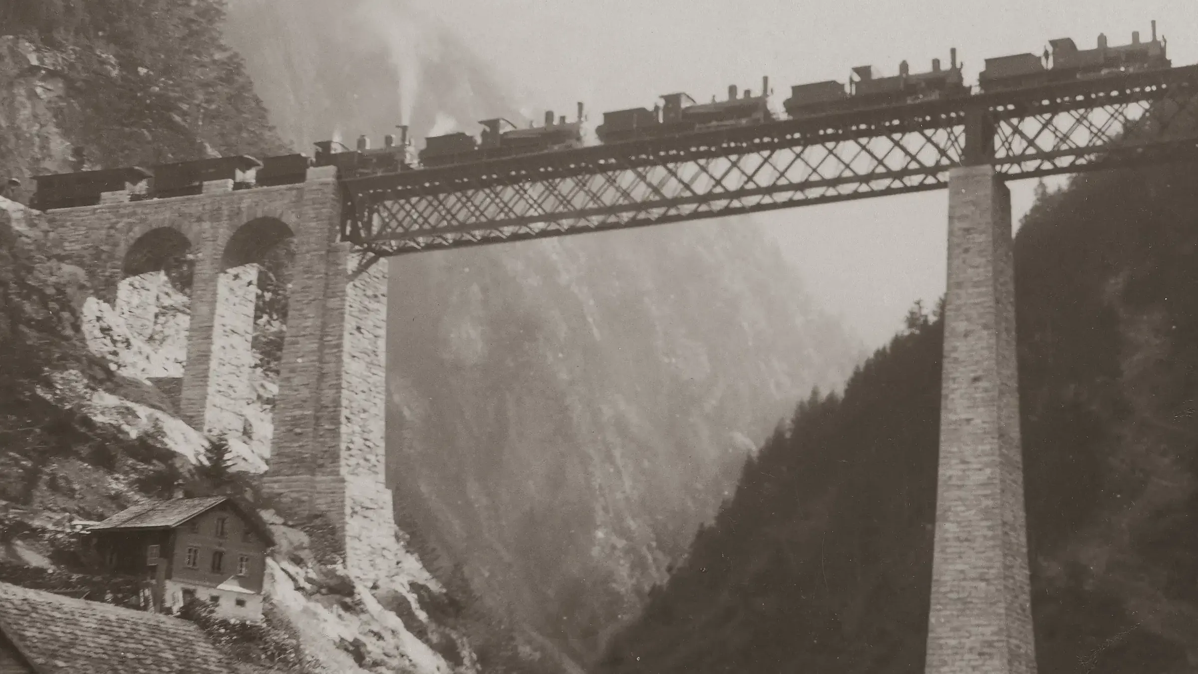 Five steam locomotives stand in a row on an iron lattice railway bridge carried on tall stone piers over a misty Alpine gorge