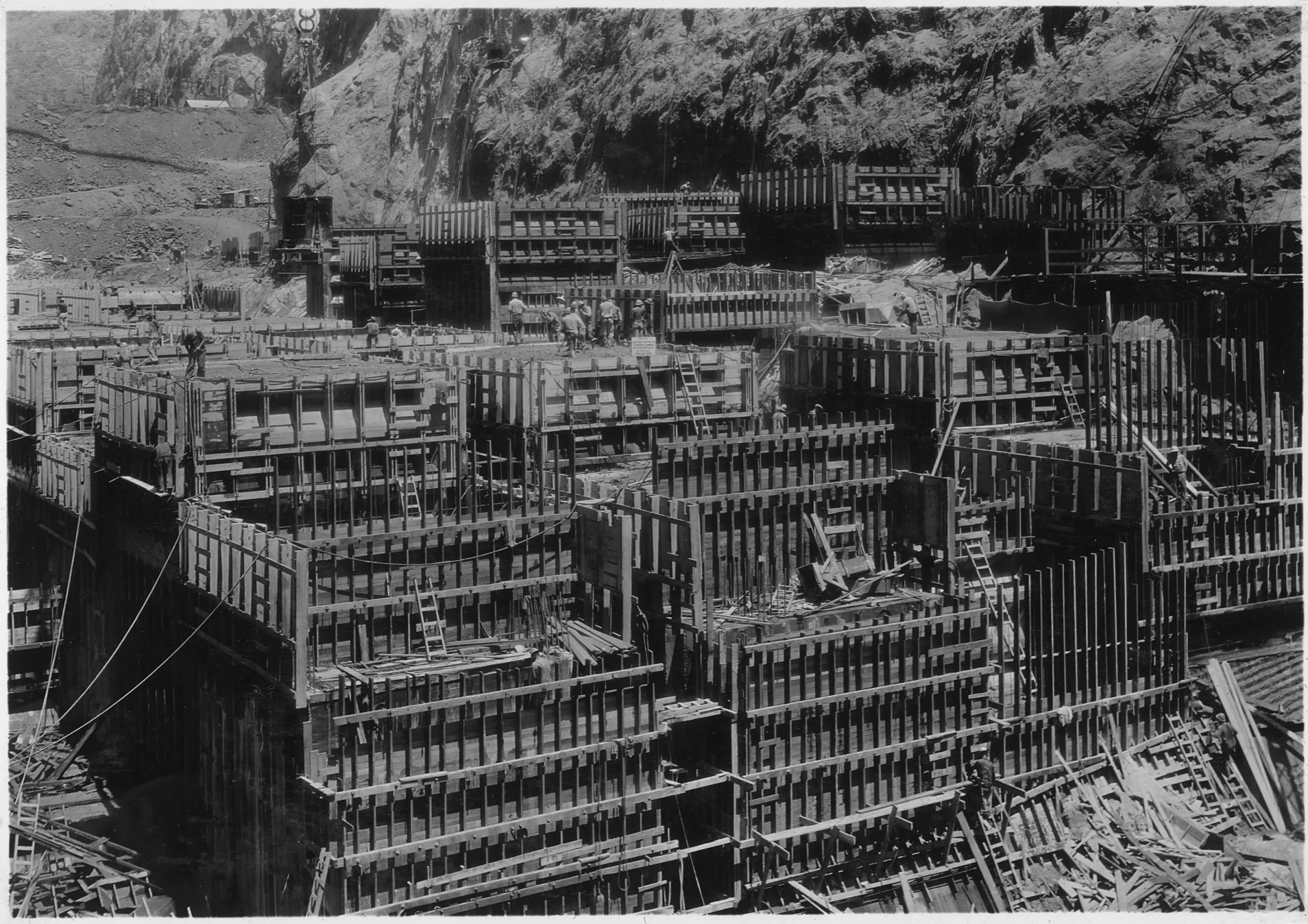 A dense field of wooden formwork boxes, ladders and scaffolding rising in stepped blocks at the base of a rocky canyon wall, with small figures of workers on top