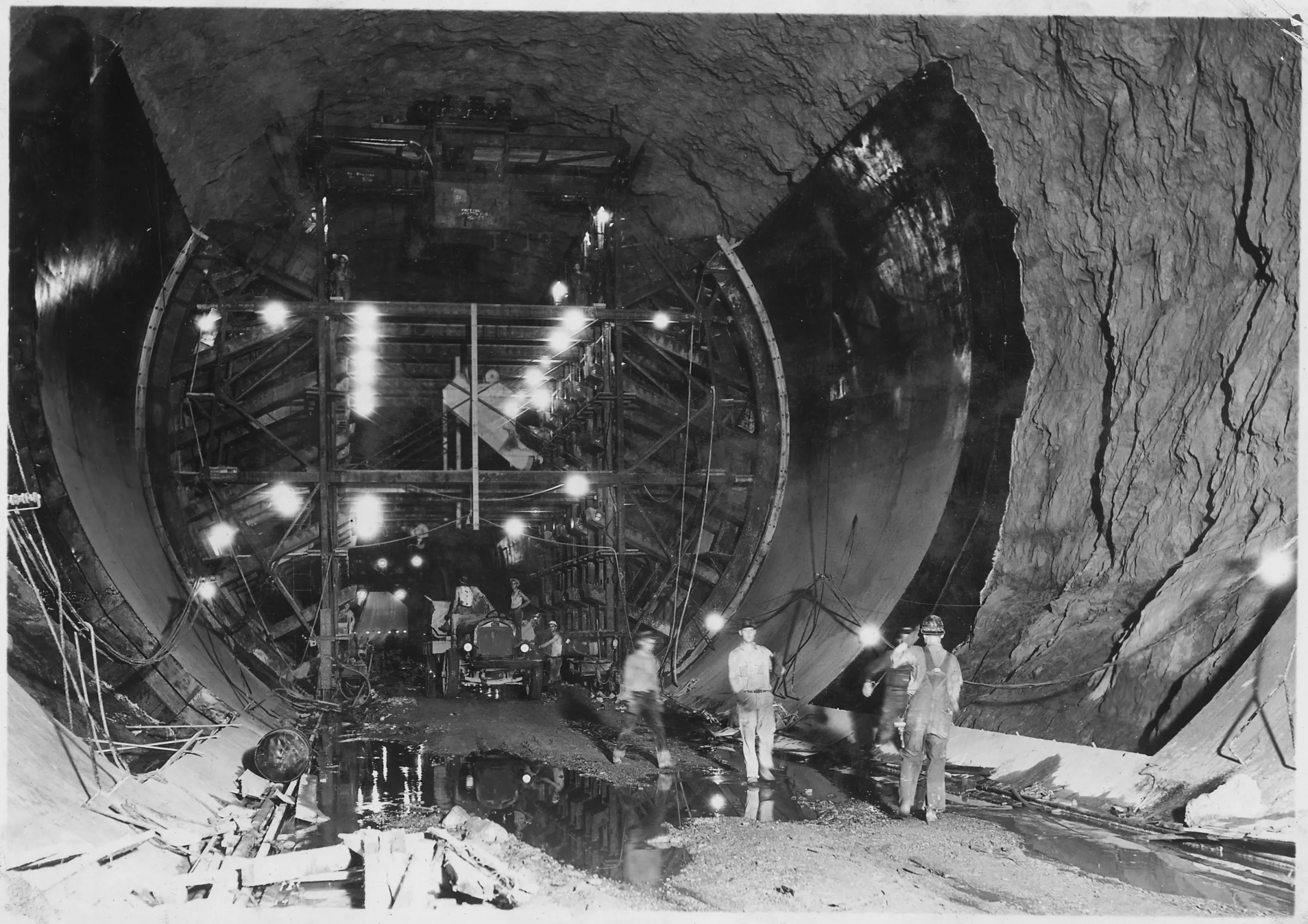 Inside a huge circular tunnel, a lit steel formwork carriage stands between a rough blasted rock wall and a smooth curved concrete lining, with workers and a truck on the wet floor