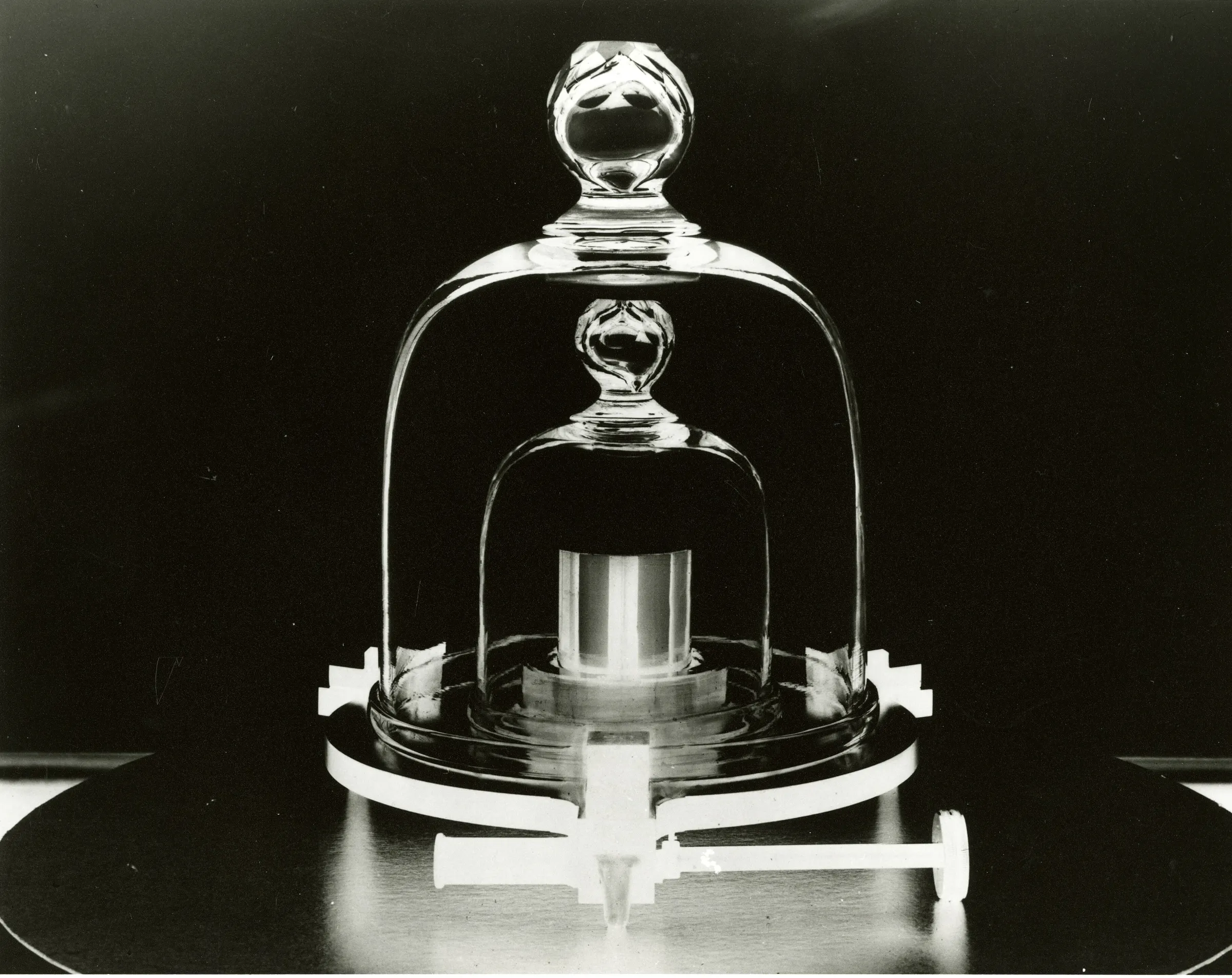 A polished metal cylinder under two nested glass bell jars on a round base, lit against a black background