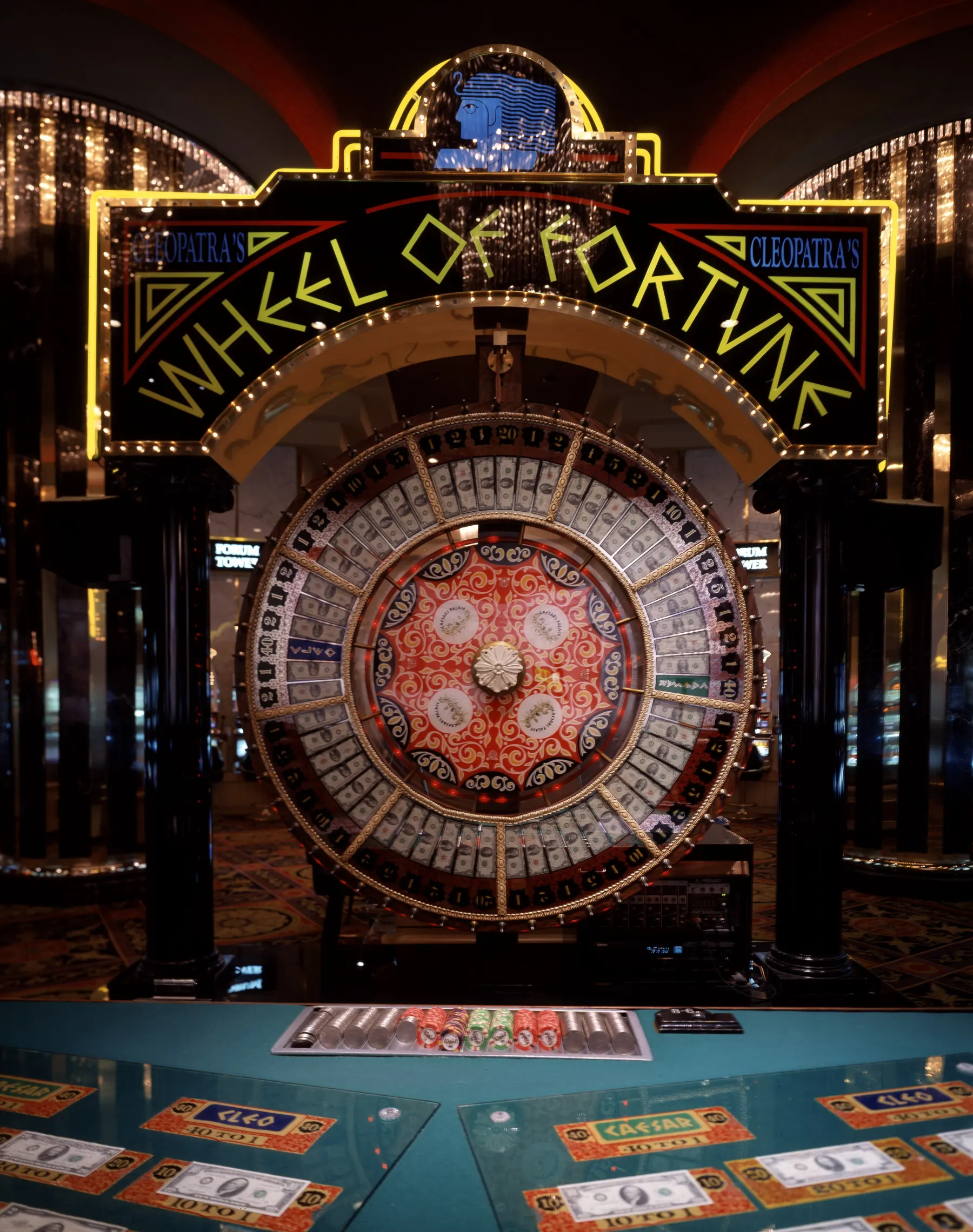 A large illuminated casino money wheel lined with dollar bills under a neon Wheel of Fortune sign, above a betting table with dollar notes under glass