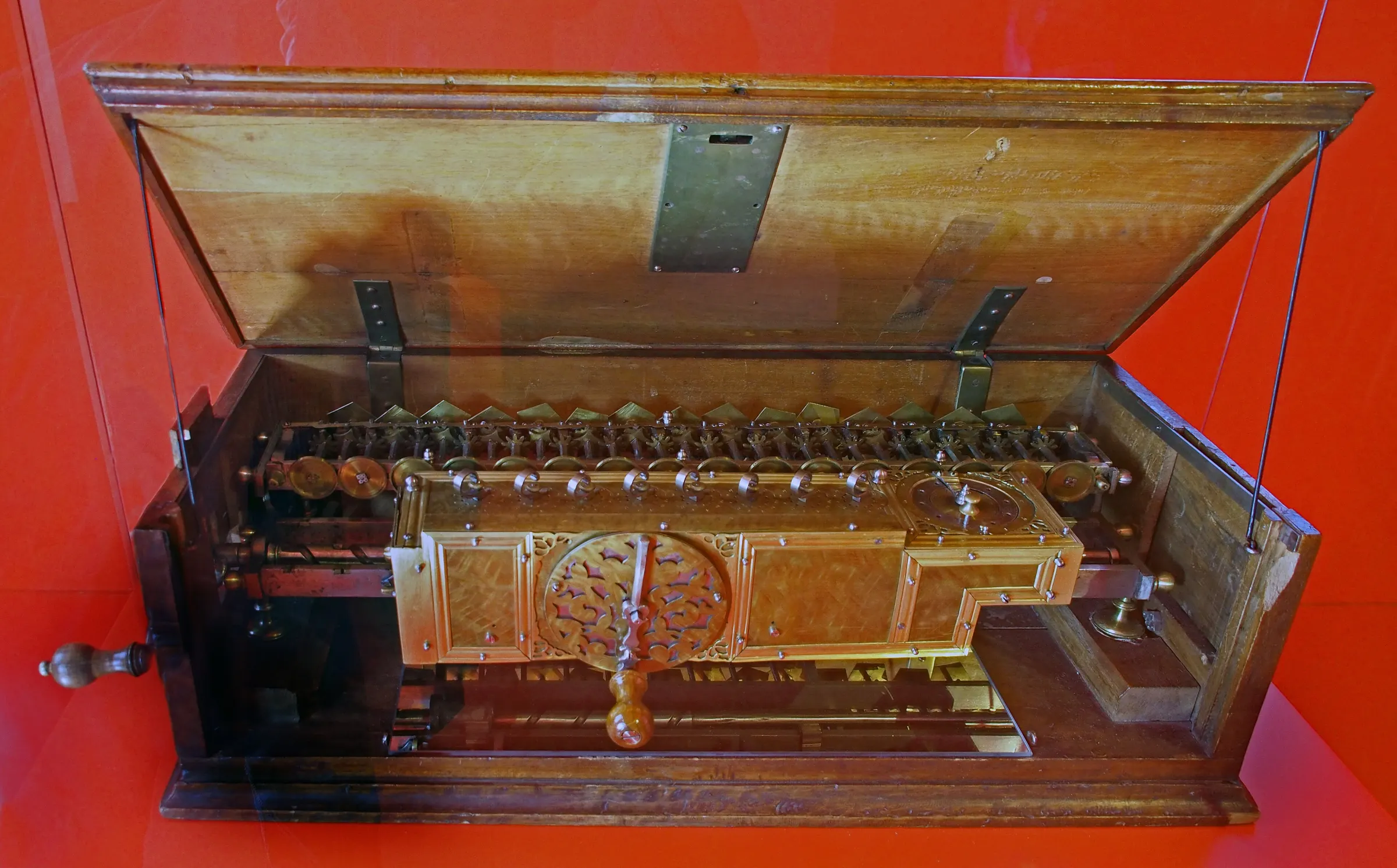 An ornate brass calculating mechanism with a row of stepped drums and a crank handle inside an open wooden case