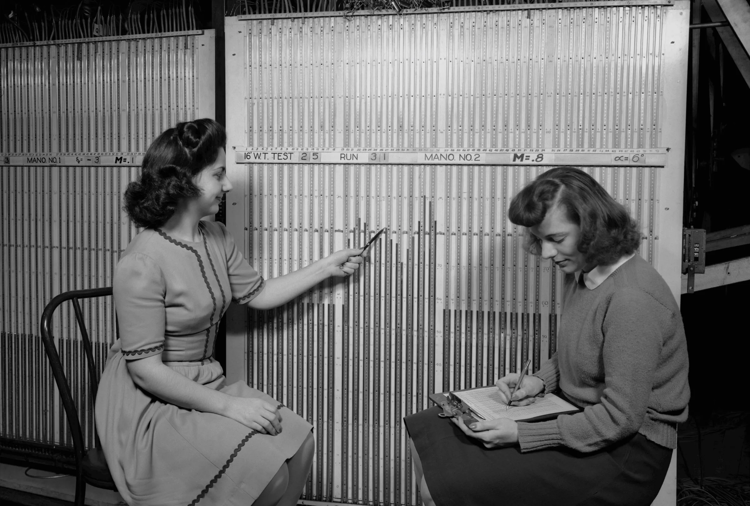 Two women sit before a wall of vertical glass manometer tubes labelled with the test and run number; one points to a column with a pencil while the other records readings on a clipboard