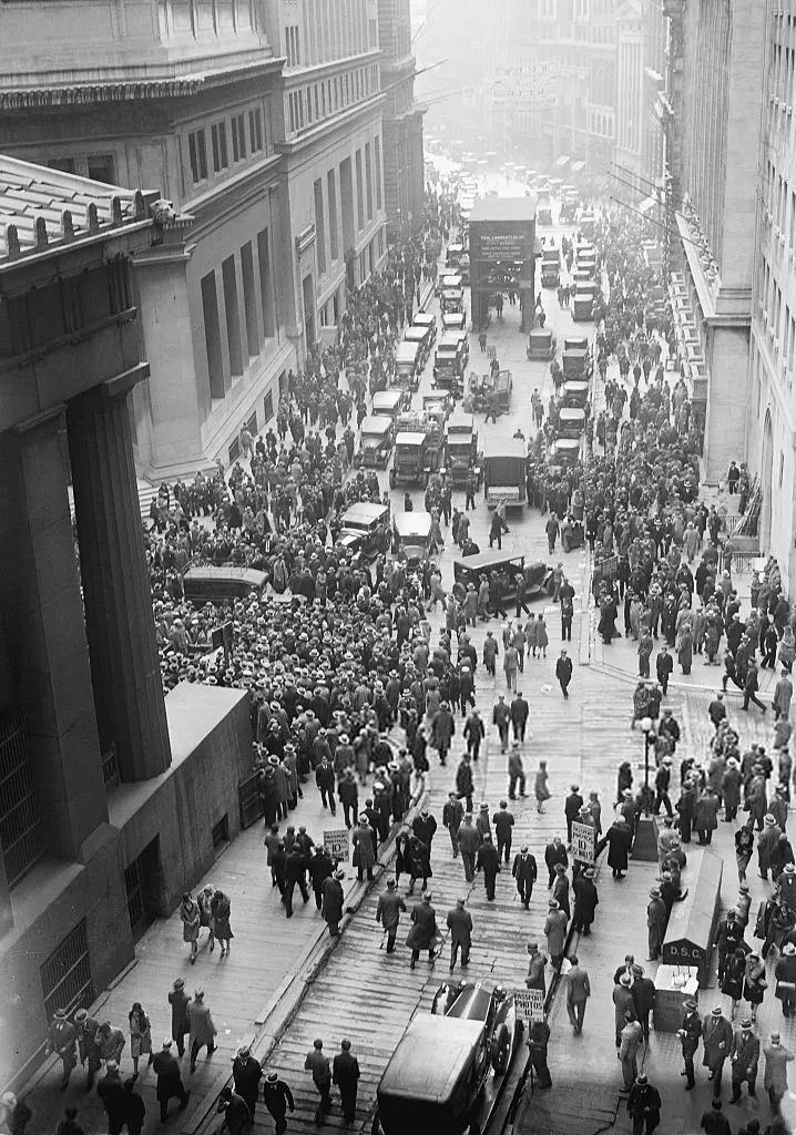 View from above of a dense crowd of men in hats filling a narrow street between tall stone buildings, with cars stuck among them