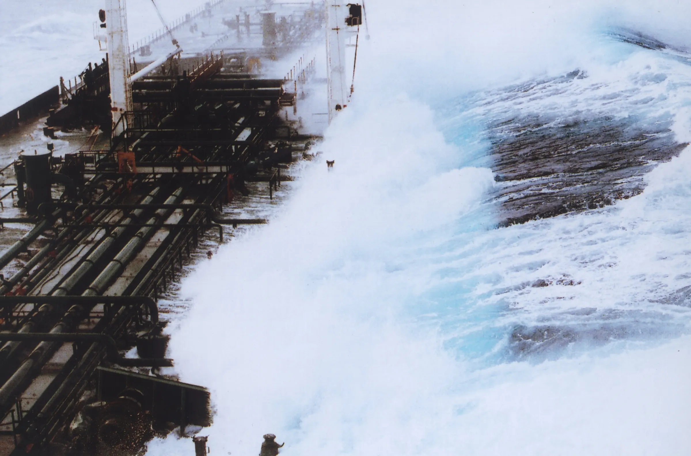 White water from a huge wave pouring across the pipe-covered deck of a tanker, with a churning blue sea beyond