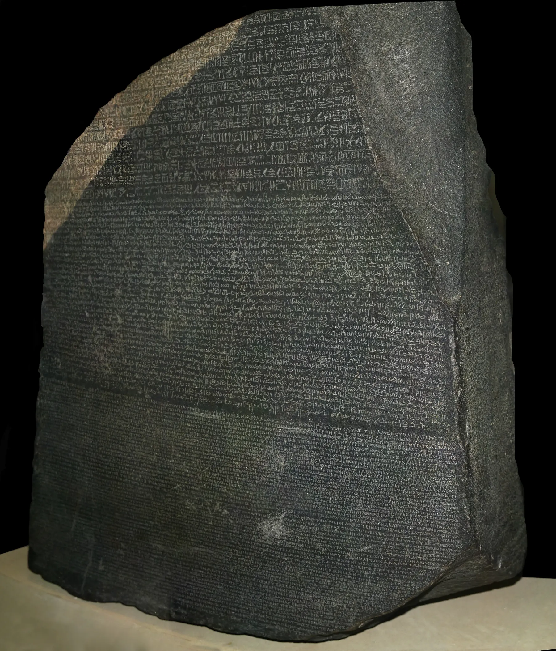 A dark grey granodiorite slab with a broken top edge, its face covered in three bands of inscriptions in hieroglyphic, Demotic and Greek scripts