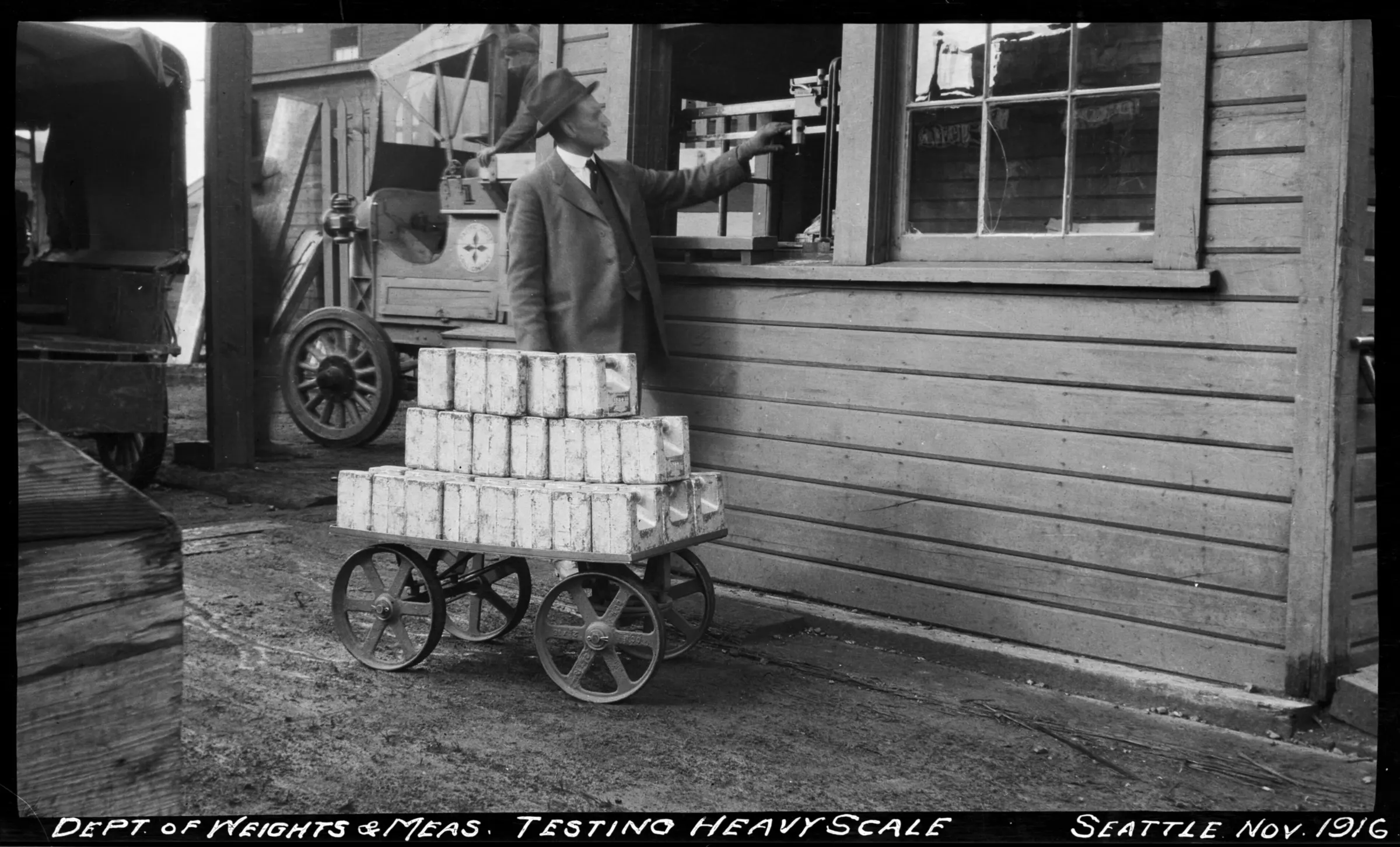 A man in a hat and overcoat adjusts the beam of a scale inside a shed window while a wheeled cart stacked with rectangular test weights stands beside him