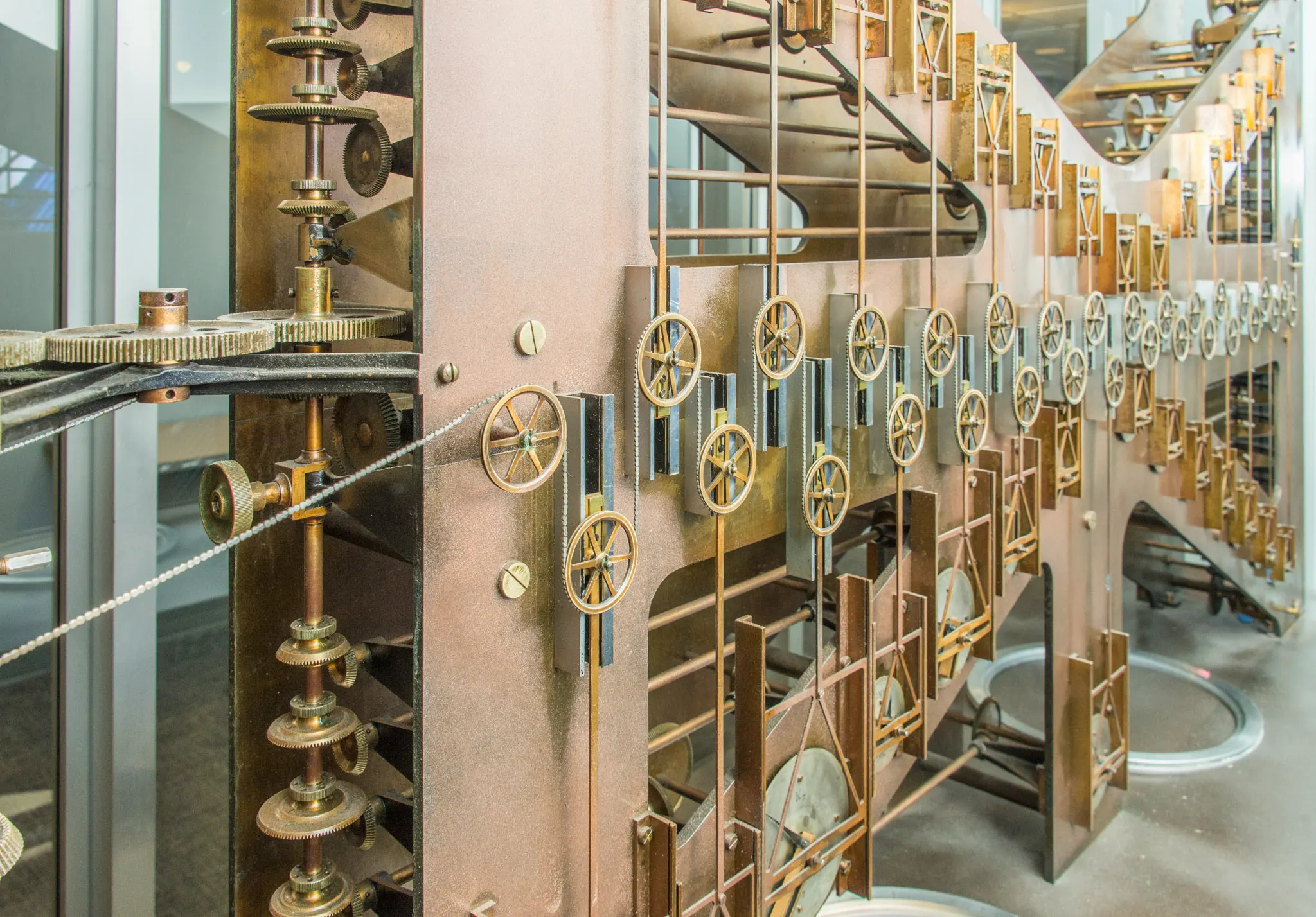 Rows of brass pulleys, gears and sliding frames on the copper-coloured side of a large mechanical analog computer, with a chain running across the wheels