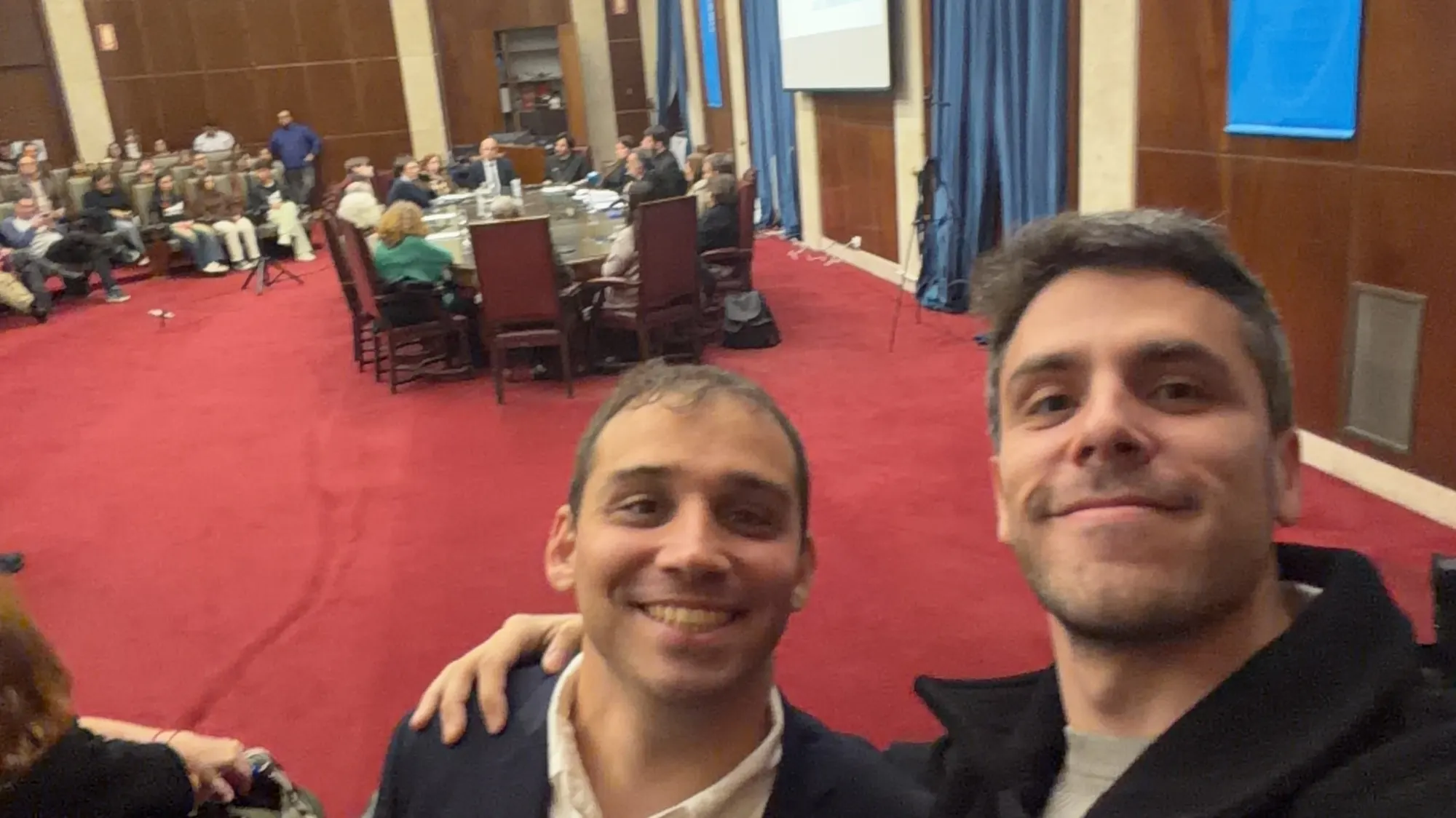 Una selfie de dos hombres sonrientes en un salón con paredes de madera y alfombra roja. Detrás, gente sentada alrededor de una mesa grande y en filas de sillas durante una sesión del consejo.