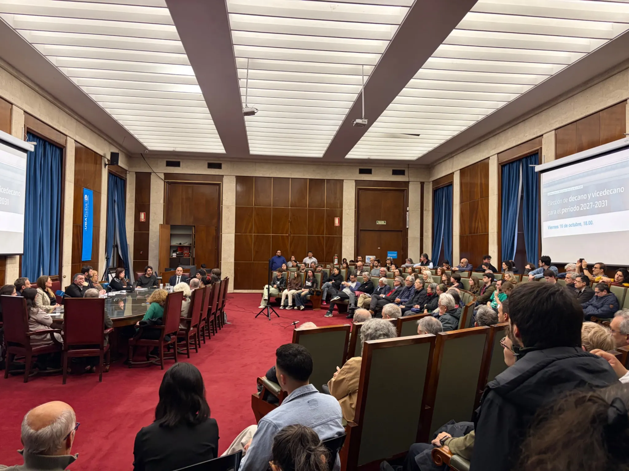 People seated around a council table and in rows of chairs in a wood-panelled hall with blue curtains and a red carpet