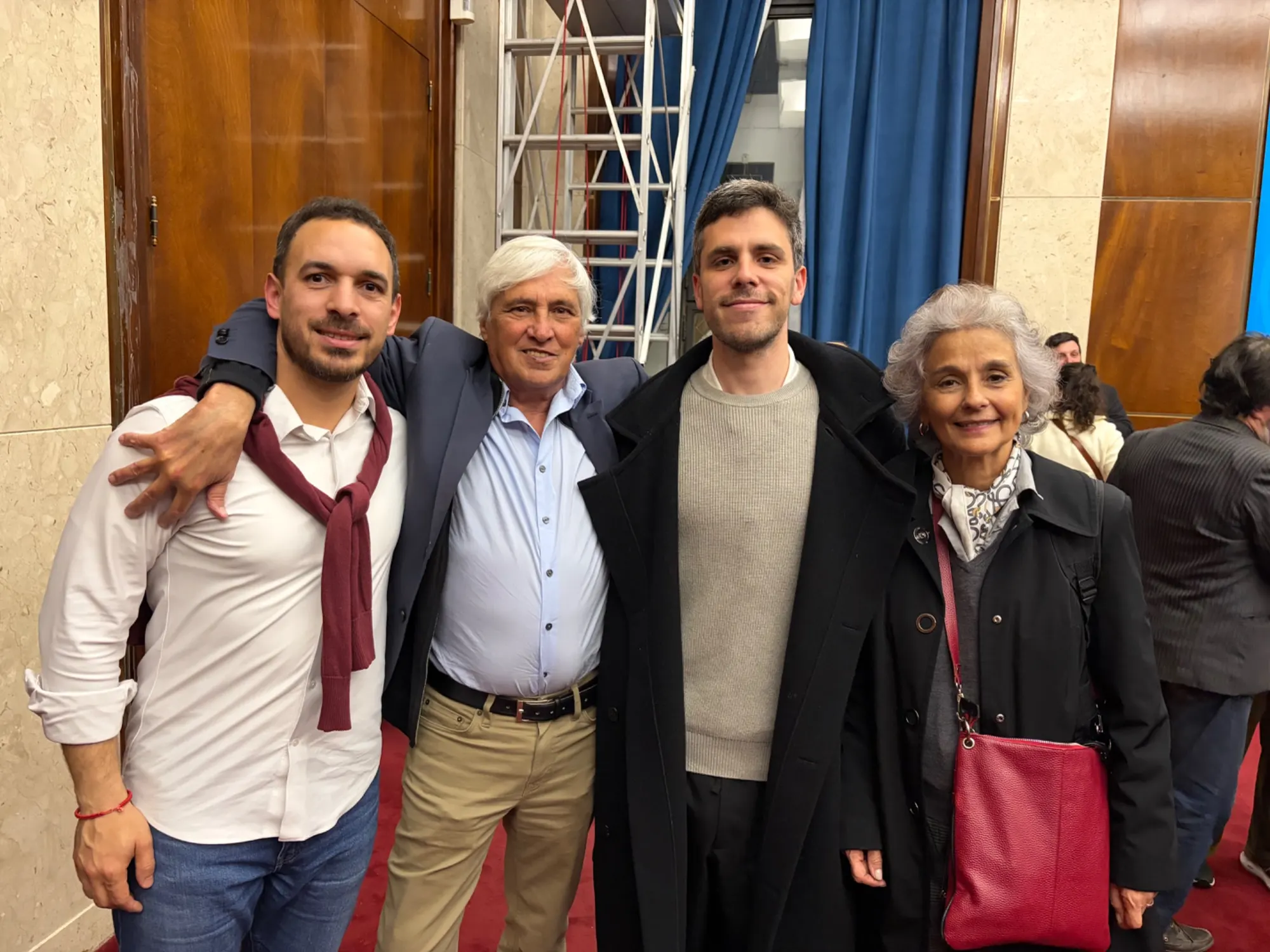 Cuatro personas posando abrazadas en un salón con paredes de madera, cortinas azules y alfombra roja, con otras personas conversando de fondo